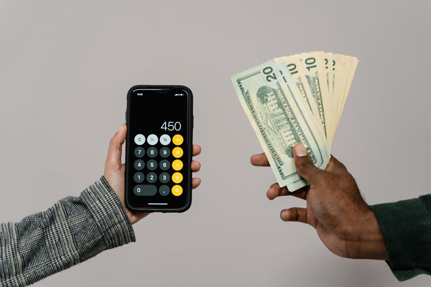 Satu tangan memegang ponsel dengan aplikasi kalkulator, berhadapan dengan tangan lain yang memegang uang tunai dolar
