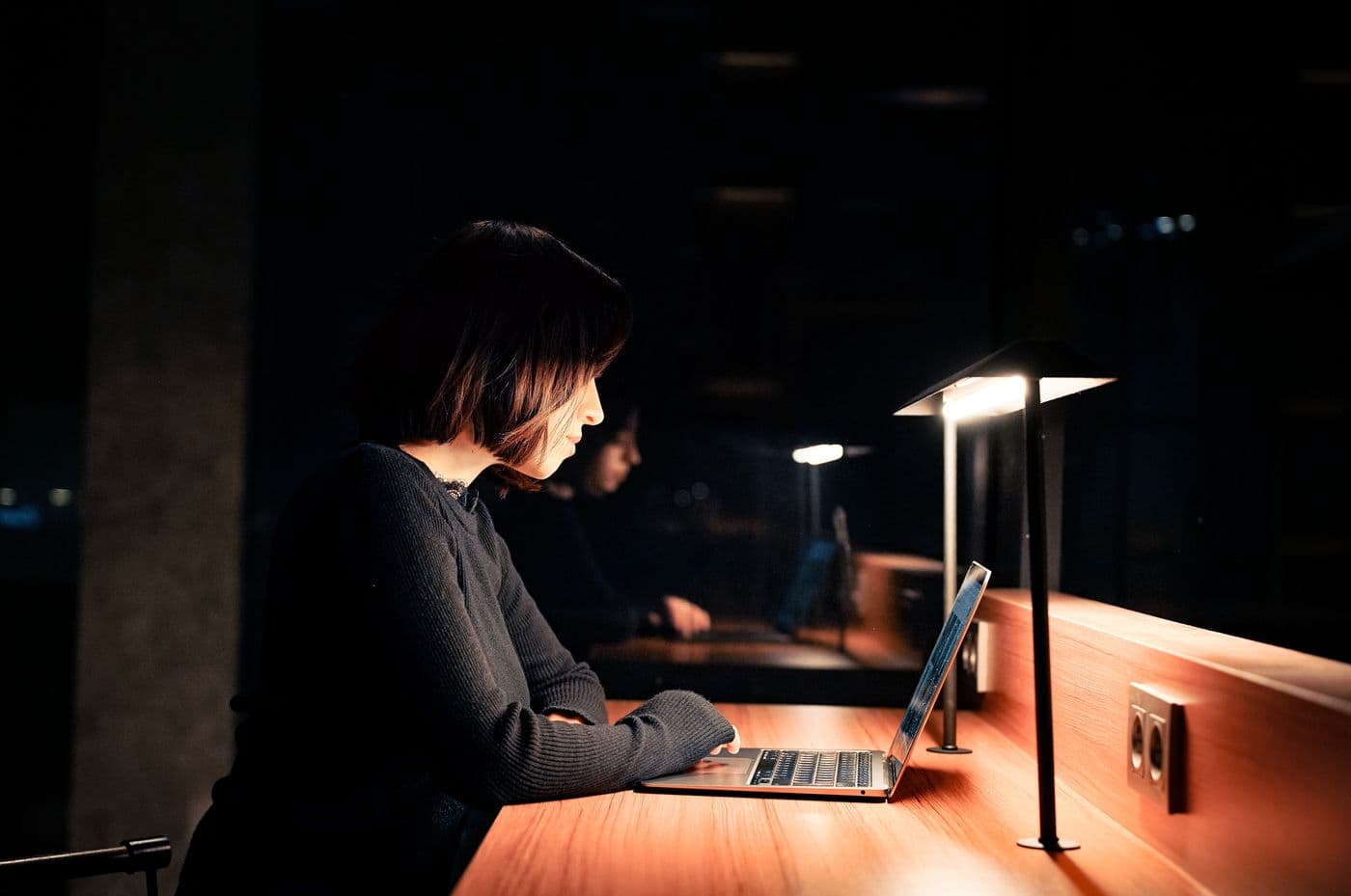 Seorang perempuan bekerja sendirian di depan laptop pada malam hari, diterangi lampu meja di ruangan gelap